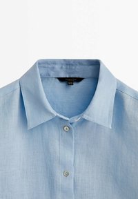 Camicia in lino azzurro chiaro con colletto classico, chiusura frontale con bottoni e una texture liscia e traspirante. Dettagli di cucitura discreti visibili.
