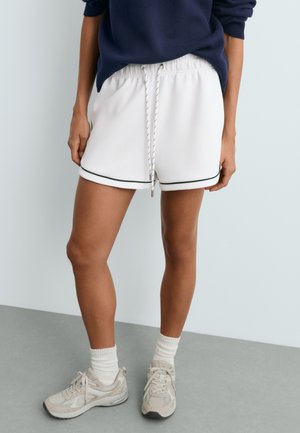 Personne portant un short blanc à cordon avec une bordure noire, un pull bleu marine, des chaussettes blanches et des baskets beiges, debout sur un sol gris.