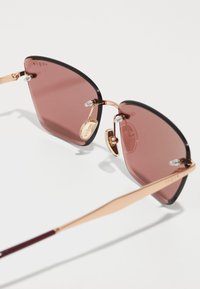 Occhiali da sole a forma di occhio di gatto con lenti rosa, bordi neri e montatura in metallo color oro. Include cuscinetti nasali trasparenti per il comfort.