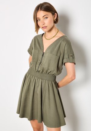 Žena s krátkými hnědými vlasy, která nosí zelené šaty s krátkými rukávy, předním zipem a nařaseným pasem, stojící před jednoduchým světlým pozadím.