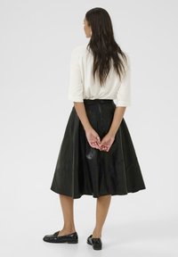 Svart imitationsläder midi kjol med en utställd design och geometriska sömmar, kombinerad med en vit topp och svarta loafers.