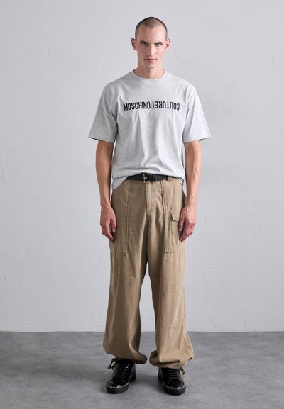 Graues Baumwoll-T-Shirt mit "MOSCHINO COUTURE!" in Schwarz bedruckt, kombiniert mit lockeren beigen Cargo-Hosen und schwarzen Schnürschuhen.