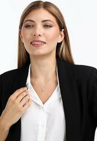 Silberne Halskette mit einem runden Anhänger, getragen über einem weißen Hemd und unter einem schwarzen Blazer. Glatte Metalloberfläche, minimalistisches Design.