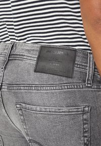 Primer plano de un bolsillo trasero de unos vaqueros de mezclilla grises y una etiqueta de piel con la marca "JACK & JONES", combinado con una camisa de rayas.