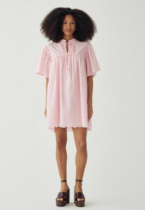 Kobieta o bujnych kręconych włosach, ubrana w luźną, jasnoróżową sukienkę z falistym dołem i ciemnobrązowe sandały na platformie, stoi na tle jednolitego tła.