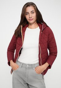 Veste matelassée bordeaux avec une capuche, fermeture éclair à l'avant et accent jaune. Portée par-dessus une chemise blanche, associée à un jean gris.