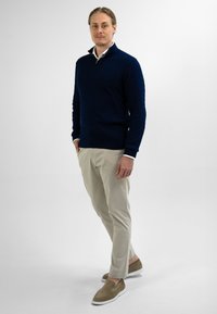 Man staat tegen een witte achtergrond, gekleed in een marineblauwe kabeltrui, beige broek en bruine instappers, met zijn handen in de zakken en een lichte glimlach.