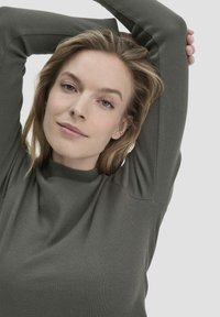 Dunkelgrünes, geripptes Langarmoberteil mit hohem Halsausschnitt und weicher Textur. Die Ärmel sind eng anliegend, und der Stoff fällt leicht an der Vorderseite.