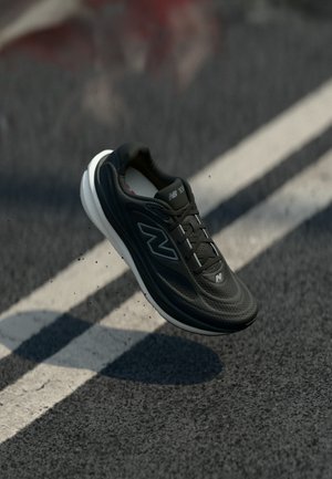 Black athletic shoe with a textured surface and white sole, featuring a lacing system and logo details, suspended above a road surface.