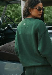 Zielona bluza o luźnym kroju, z haftowanym napisem „self.” na tylnej stronie kołnierza. Gładka faktura, prosty design.