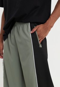 Athletic pants with a black and olive green design, featuring a zippered pocket and white side stripes. Made from smooth, lightweight fabric.