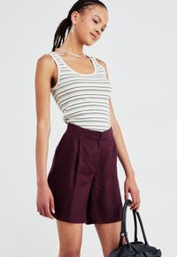 Linen high-waisted shorts in deep plum, featuring a front button and pleat design. Paired with a fitted, striped tank top.
