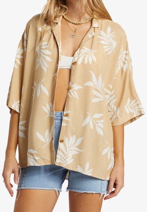 Kobieta ubrana w beżową koszulę na krótki rękaw z białym liściastym nadrukiem, na białym topie bandeau oraz jasnoniebieskich szortach jeansowych.