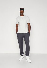 T-shirt branca ajustada com pequeno logótipo, combinada com calças de treino cinzentas com punhos às riscas e ténis brancos. Usa uma faixa preta na cabeça.