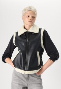 Melna ādas veste ar krēmkrāsas apdarītiem jakas apkakli un sānu paneļiem, ar priekšējo rāvējslēdzēju un divām apakšējām kabatām.