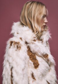 Cappotto in pelliccia sintetica con un mix di motivi bianchi e marroni, con colletto alto e superficie testurizzata e soffice. Vista laterale.