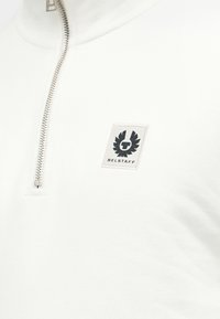 Λευκό ένδυμα με φερμουάρ στο ένα τέταρτο, ασημένιο φερμουάρ και επιγραφή με λογότυπο Belstaff που απεικονίζει ένα μαύρο φτερωτό έμβλημα και κείμενο στο στήθος.