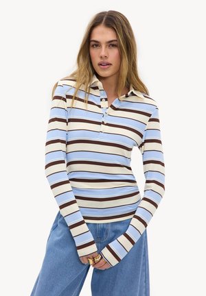 Jonge vrouw die een poloshirt met lange mouwen en een kraag draagt, met blauwe, bruine en witte horizontale strepen, gecombineerd met losse blauwe jeans en ringen aan haar vingers.