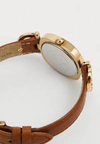 Goldfarbener Uhrengehäuse mit runder Krone, braunem Lederarmband und Schnalle, von hinten auf einem weißen Hintergrund dargestellt.