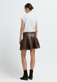 Jupe en cuir synthétique brun avec un design évasé et à étages, dotée d'une taille ajustée et de coutures visibles. Portée avec une chemise blanche à col et des bottes noires à la cheville.