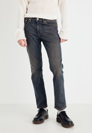 Person trägt ausgewaschene schwarze Jeans mit geradem Bein, schwarze Schnürschuhe, hellgraue Socken und einen weißen Strickpullover mit langen Ärmeln.