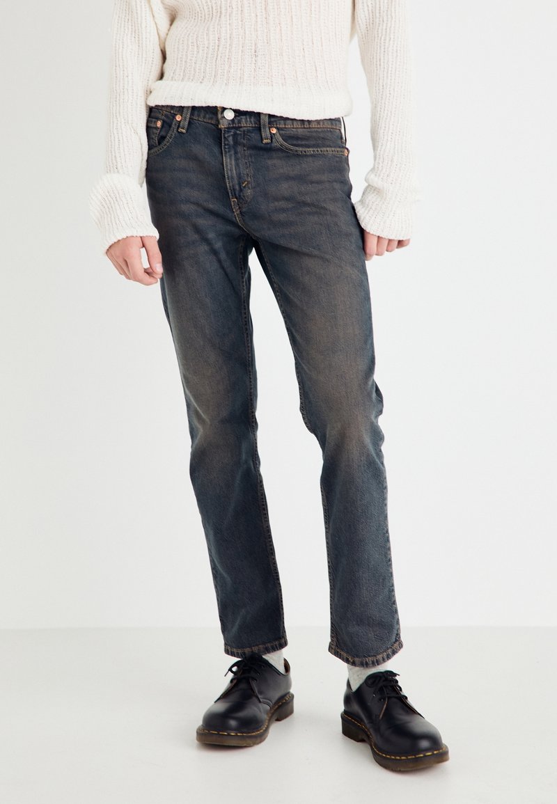 Personne portant un jean droit noir délavé, des chaussures noires à lacets, des chaussettes gris clair et un pull en maille blanc à manches longues.