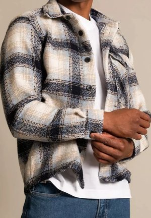 Mężczyzna poprawiający mankiet beżowej, czarnej i niebieskiej koszuli polarowej w kratę na białej koszulce i niebieskich dżinsach.