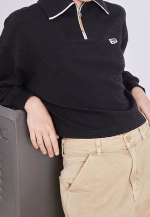 Black textured pullover with a high collar, zipper, and white trim, paired with beige pants featuring side pockets and a button closure.