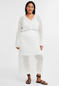 Weißes Häkel-Zweiteiler-Set mit tiefem V-Ausschnitt, langärmliger Strickjacke und passendem Rock, beide mit filigranen Spitzenmustern und offenen Strickdetails.