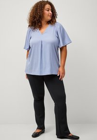Light blue, short-sleeve top with a V-neck and pleated front, paired with black slim-fit pants and flat black shoes on a neutral backdrop.