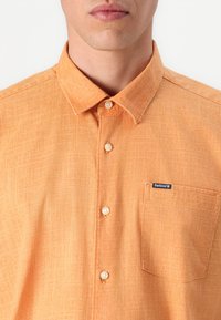Camisa laranja abotoada, feita de tecido texturizado, com um bolso no peito e um pequeno rótulo da marca no bolso.