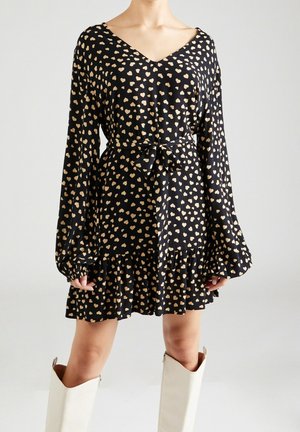 Schwarzes Kleid mit V-Ausschnitt, langen Ärmeln und goldenem Herzmuster. Mit Bindegürtel in der Taille und ausgestelltem Volant-Saum. Dazu weiße kniehohe Stiefel getragen.