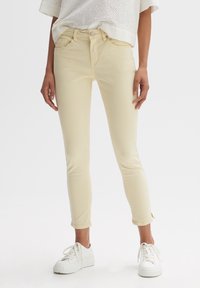 Jean jaune clair à coupe ajustée, avec deux poches avant et ourlets retroussés, associé à des baskets blanches. Texture lisse et détails de couture subtils.