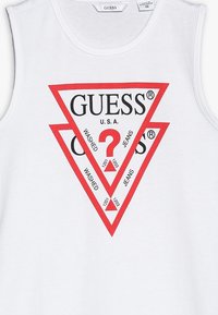 Biała koszulka bez rękawów z czerwonym trójkątnym logo z napisem „GUESS U.S.A.” oraz tekstem „WASHED JEANS” w pogrubionej czarnej czcionce.