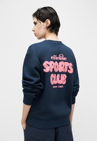 Άτομο με κοντά σκούρα μαλλιά που φοράει μπλε μπλούζα φούτερ με ροζ κείμενο "ellesse SPORTS CLUB since 1959" στην πλάτη, κοιτώντας πλάγια.