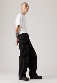 Mand med tatoveringer iført en hvid t-shirt, sorte cargo-bukser og sorte sko står sidelæns mod en ensfarvet lys baggrund.
