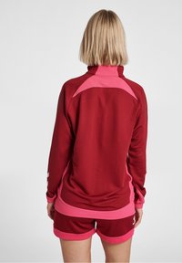Camiseta deportiva de manga larga color granate con detalles en rosa, con cuello alto y paneles de malla. Combinada con pantalones cortos granates con ribete rosa.