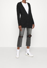 Persona vestida con un cárdigan negro abotonado, camiseta blanca, jeans grises rasgados, calcetines blancos y zapatillas blancas con logo negro.