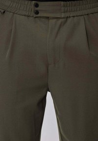 Pantalon vert olive à texture lisse, doté d'une ceinture élastique et de deux poches avant, agrémenté de boutons noirs.