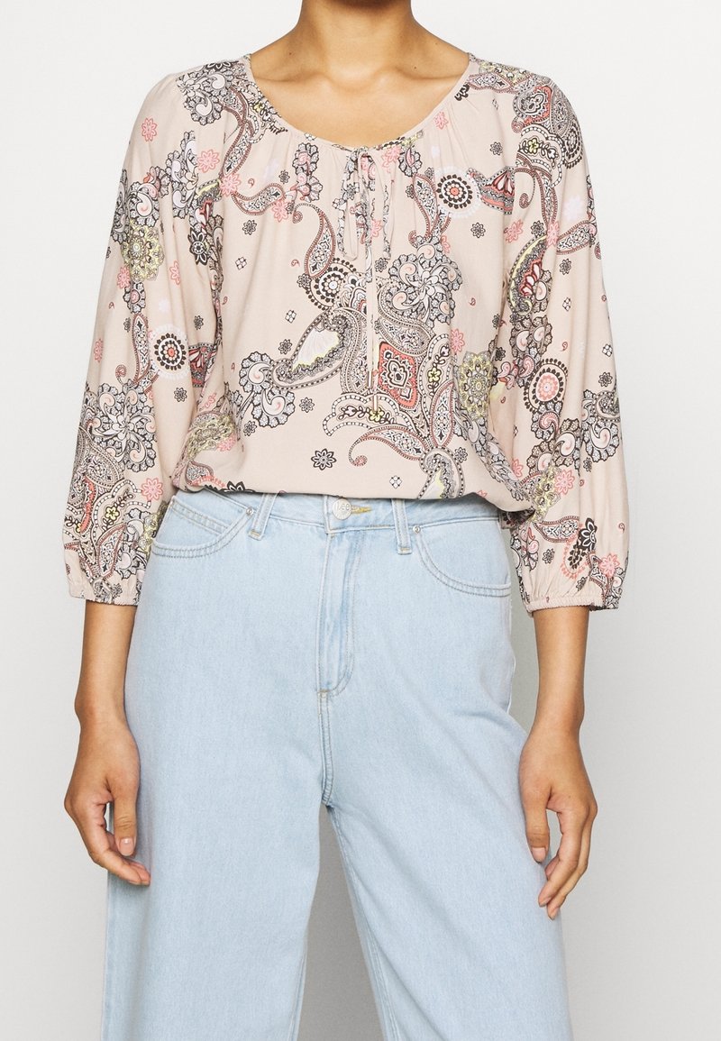 Femme portant un chemisier rose clair à motif paisley avec des manches trois-quarts, rentré dans un jean taille haute bleu clair, mains détendues sur les côtés.