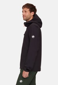 Black waterproof jacket with a hood, featuring a chest pocket, elastic cuffs, and a logo on the sleeve. Model wears green trousers.