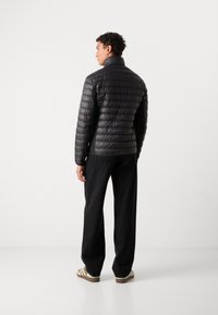 Sort pufferjakke med høj krave, quiltet design og slim fit. Modellen bærer sorte bukser og stribede sko, set fra ryggen.