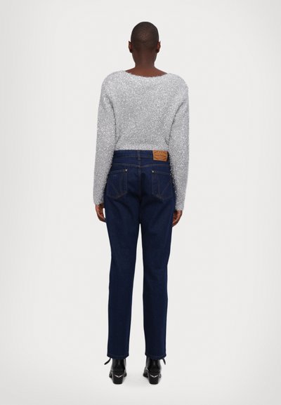 Suéter corto plateado brillante con mangas largas, combinado con jeans de mezclilla azul oscuro que presentan una etiqueta marrón y detalles en el bolsillo trasero.
