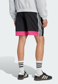 Pantalones cortos deportivos negros con un dobladillo rosa, que presentan tres rayas verticales blancas en los laterales. Se combinan con zapatos negros y calcetines a rayas blancas.
