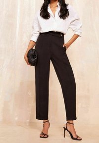 Pantalones negros de talle alto con un ajuste entallado, con cierre de botones y costuras visibles. Combinados con una camisa blanca y un bolso de mano negro.