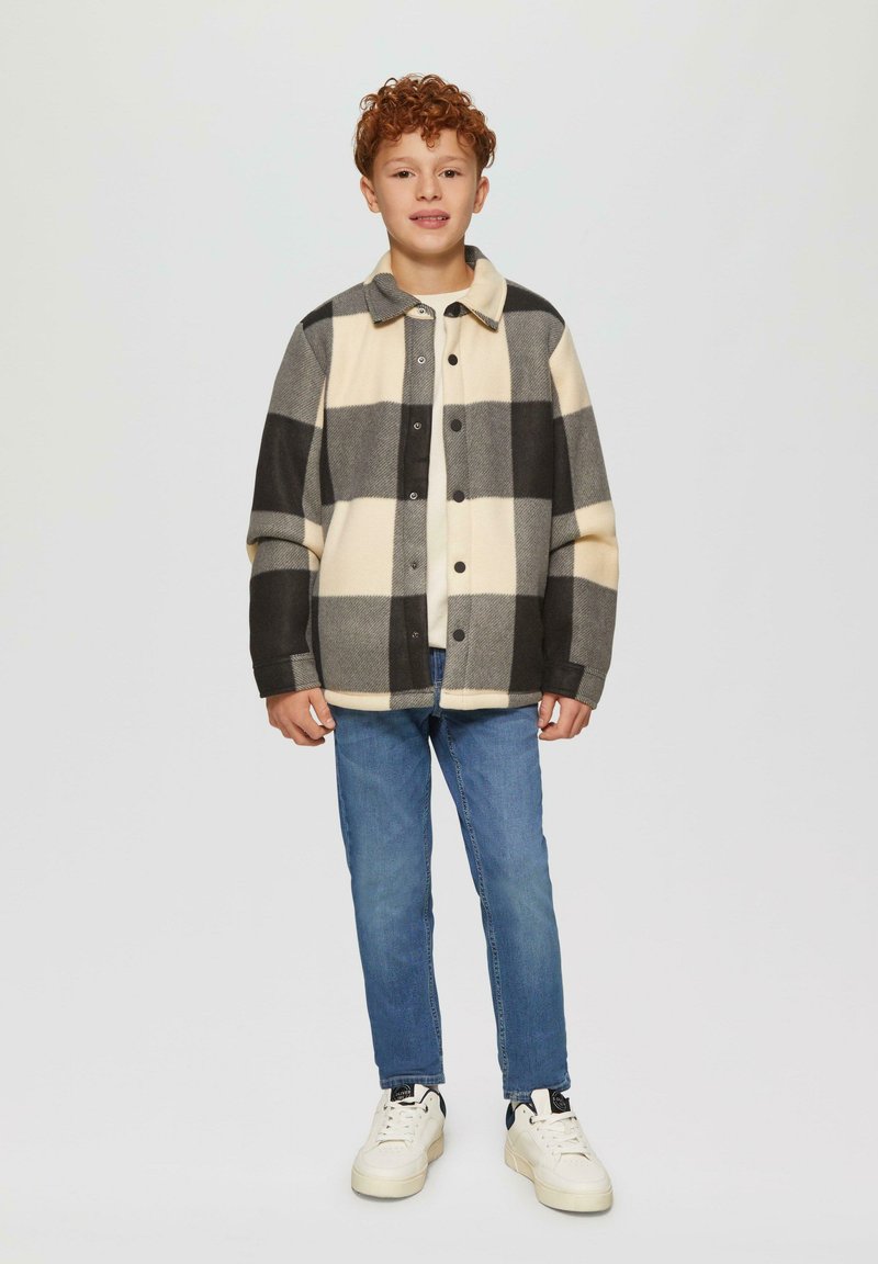 Kurtka z włóczki w kratę w odcieniach kremowym, czarnym i szarym; z guzikami z przodu i kołnierzem; noszona z niebieskimi dżinsami oraz białymi trampkami.