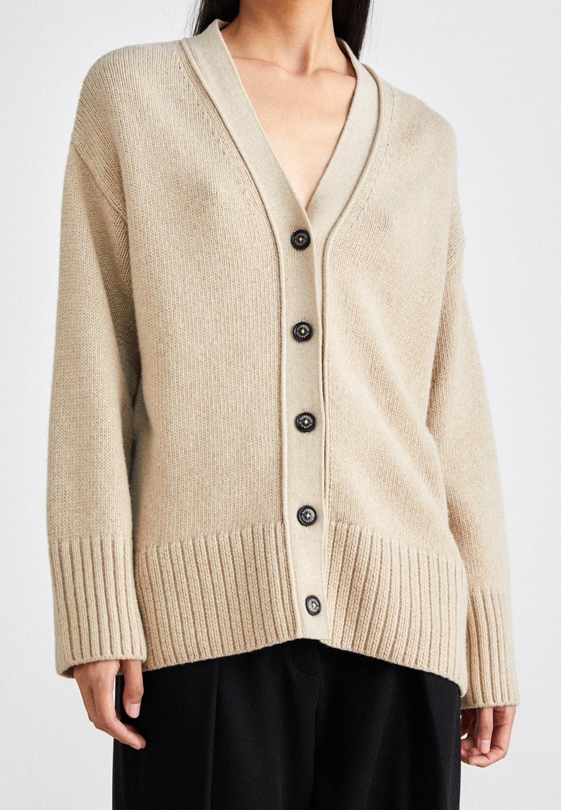 Bežový pletený cardigan s hlubokým V-výstřihem, šesti černými knoflíky a žebrovanými manžetami a spodním lemem. Měkká textura a uvolněný střih.
