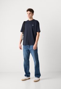 Tricou bleumarin cu mânecă scurtă și un logo mic, asortat cu blugi deschiși la culoare. Persoana poartă o geantă gri de umăr și adidași albi.