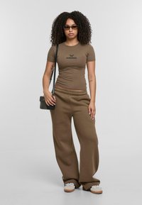 Braun enges T-Shirt mit "Follilous"-Logo, kombiniert mit lockeren braunen Sweatpants. Die Accessoires umfassen eine kleine schwarze Tasche und Sonnenbrille. Lässiger Stil.