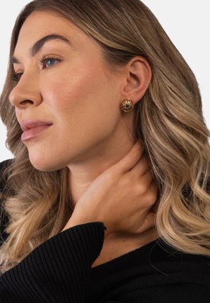 Frau mit blonden, welligen Haaren, die ein schwarzes Oberteil und goldene, runde Steckerohrringe trägt, berührt sanft ihren Hals und schaut nachdenklich zur Seite.